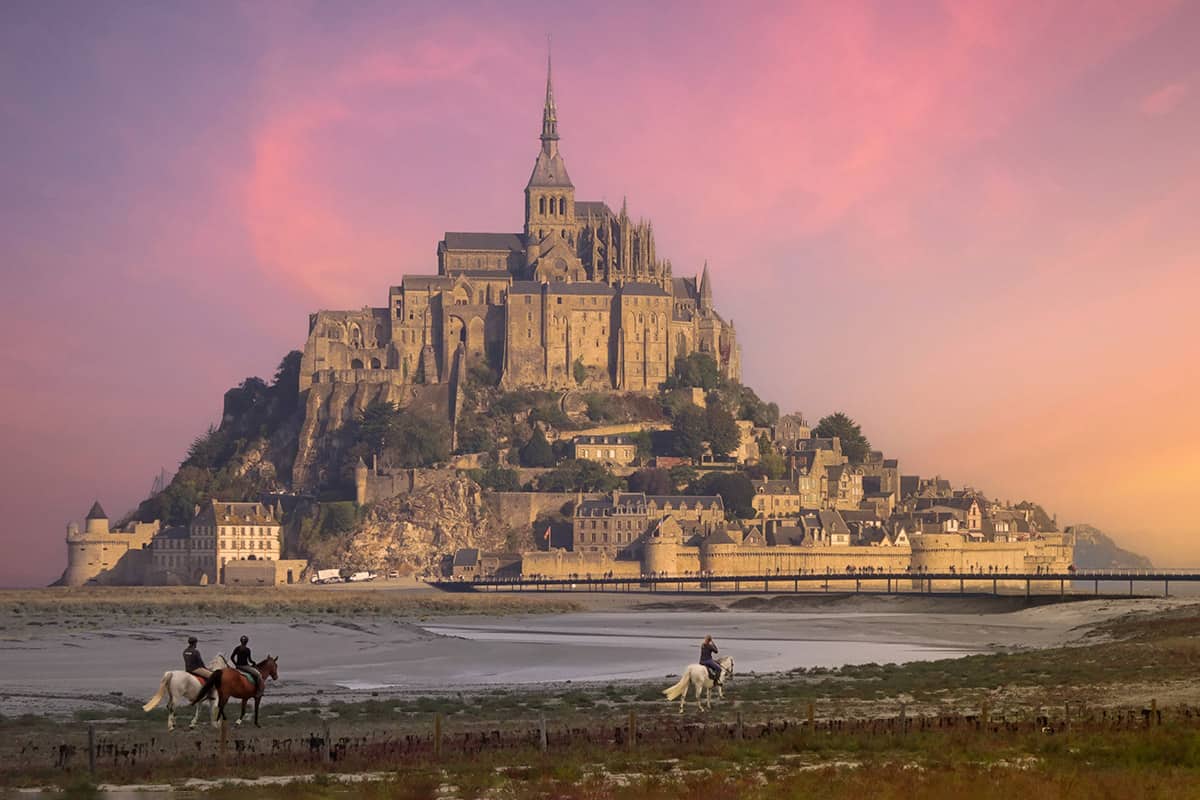 Two riders on horses looking at an island castle at sunset