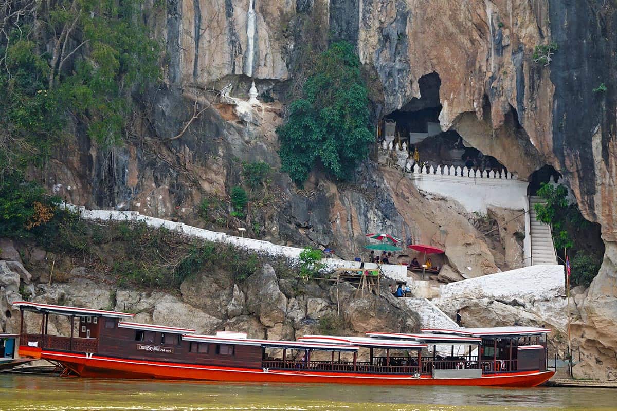 A long boat moored by the side of a river with caves in the cliff above